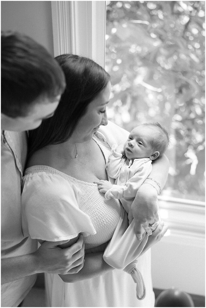 mother and father smiling at baby while standing in front of a window