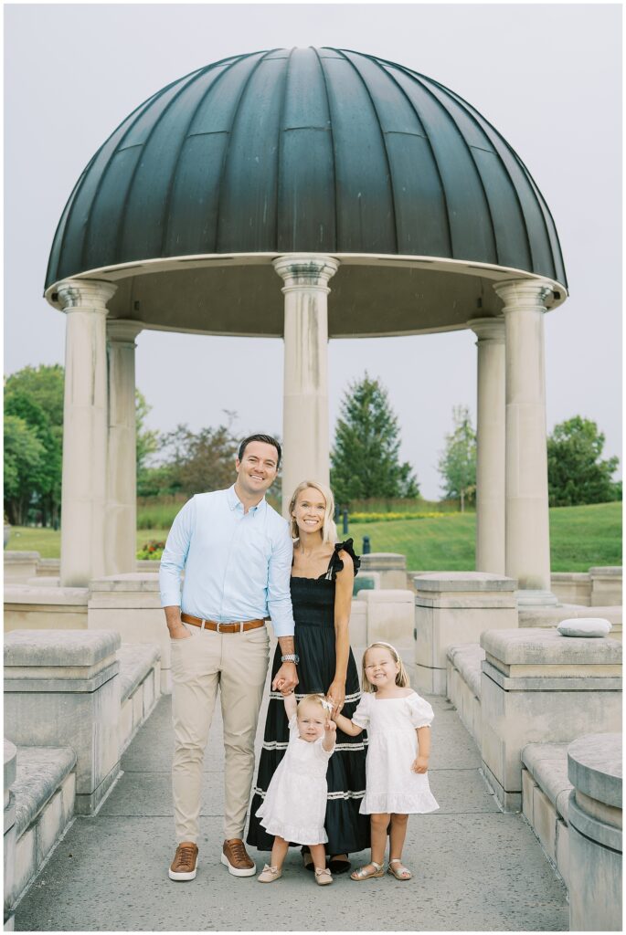Couple with two daughters in white stand in front of dome at Coxhall Gardens in Carmel Indiana