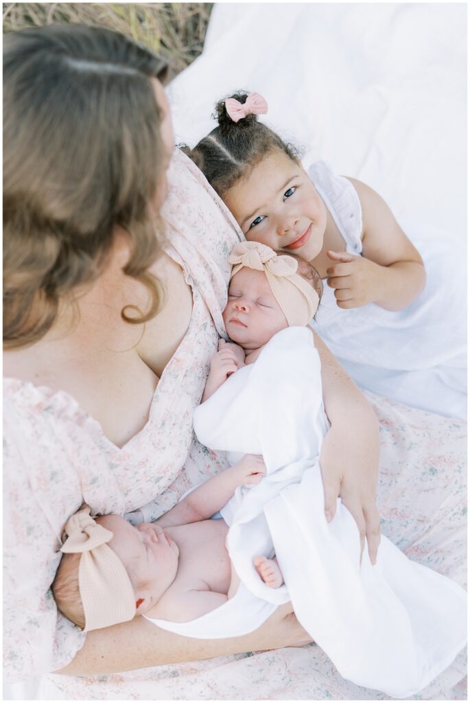 Mother in light pink holds twin newborn daughters with toddler daughter during Carmel Indiana photo session